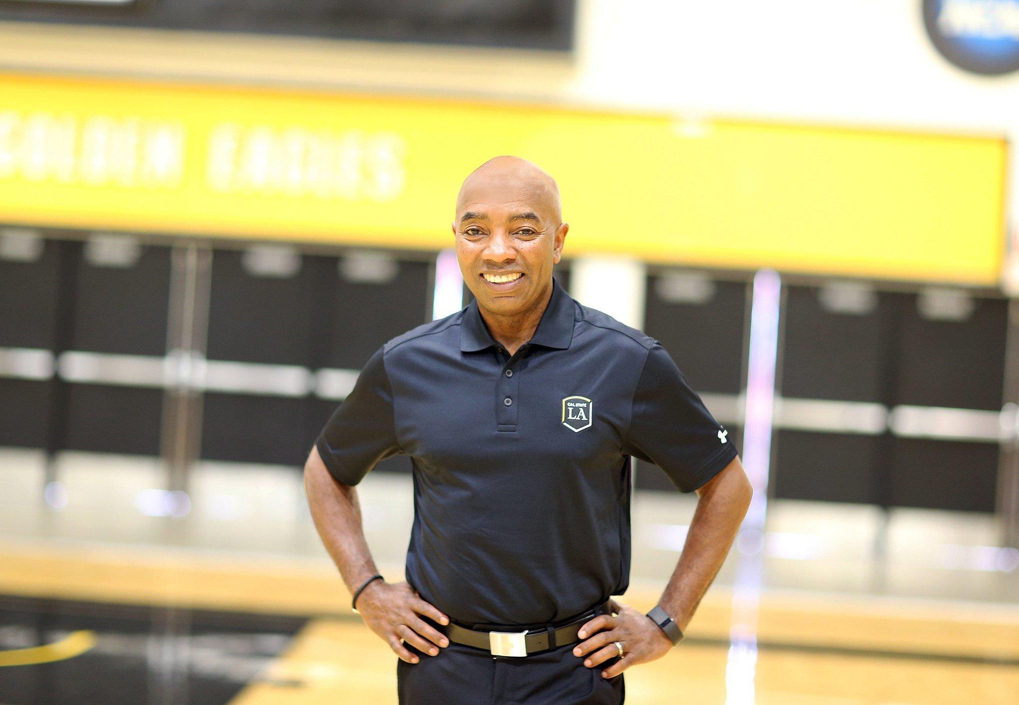 A coach in a Cal State LA polo stands in a gym with his hands on his hips.